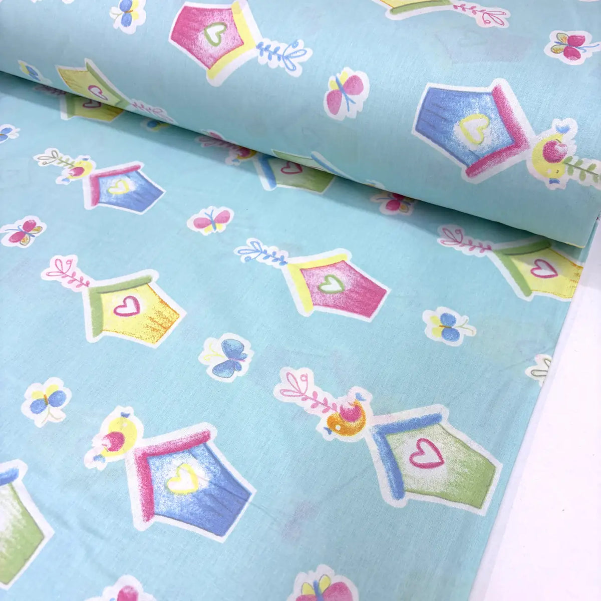 Cotton poplin fabric with colourful birdhouse, butterfly, bird and heart pattern on a light blue background.