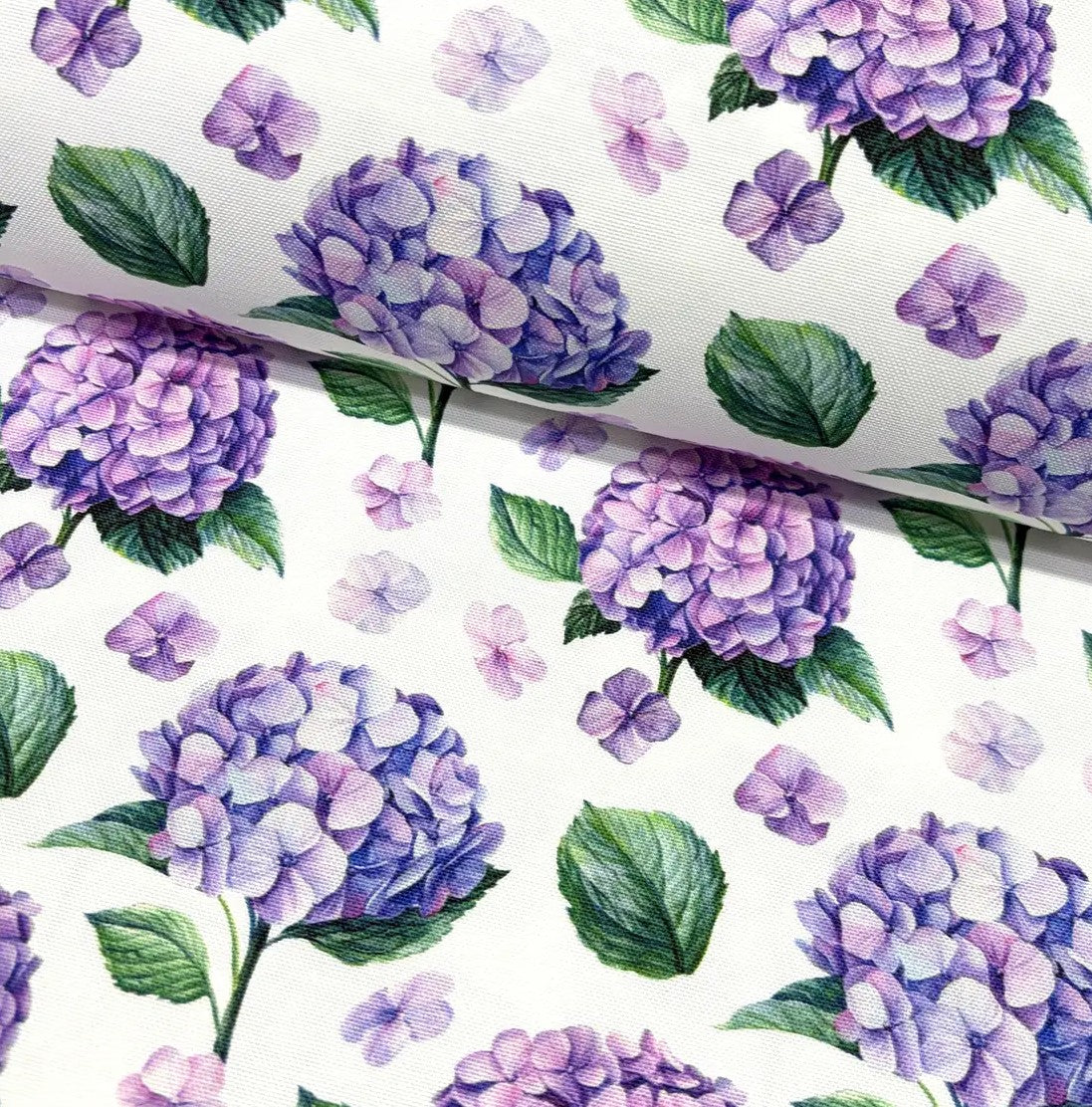 Fabric patterned with lilac hydrangea flowers and green leaves on a white background.