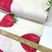 A close-up of a roll of white fabric printed with large red strawberries and green leaves, with a wooden ruler laid across the fabric to show scale.