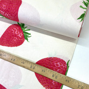 A close-up of a roll of white fabric printed with large red strawberries and green leaves, with a wooden ruler laid across the fabric to show scale.