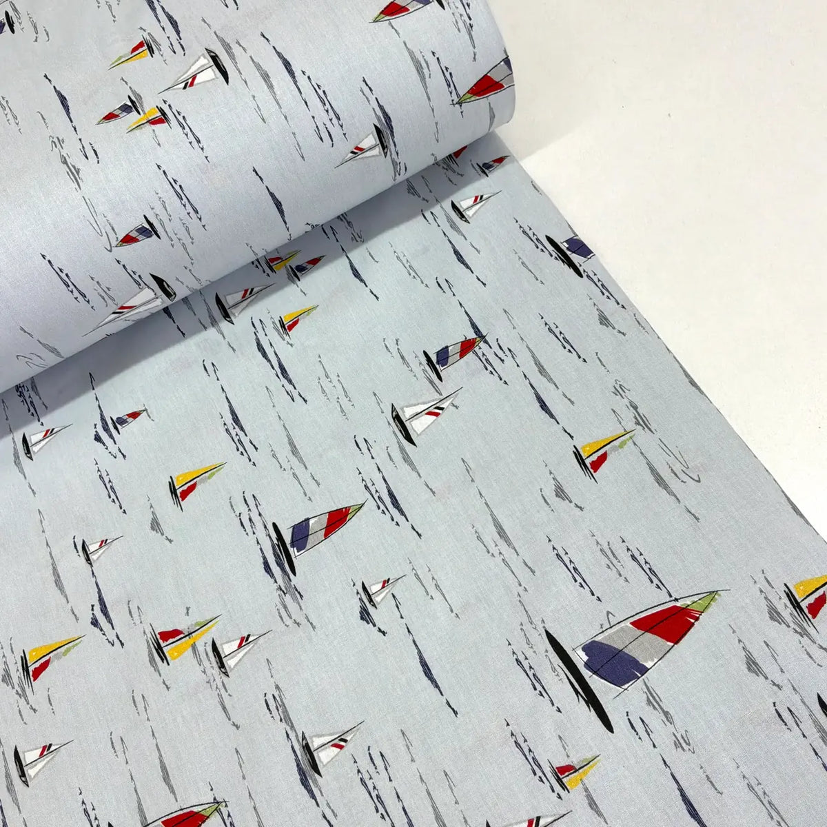 A roll of light gray fabric with a repeating print of small sailboats in red, yellow, blue, and white, alongside abstract black brushstroke lines.