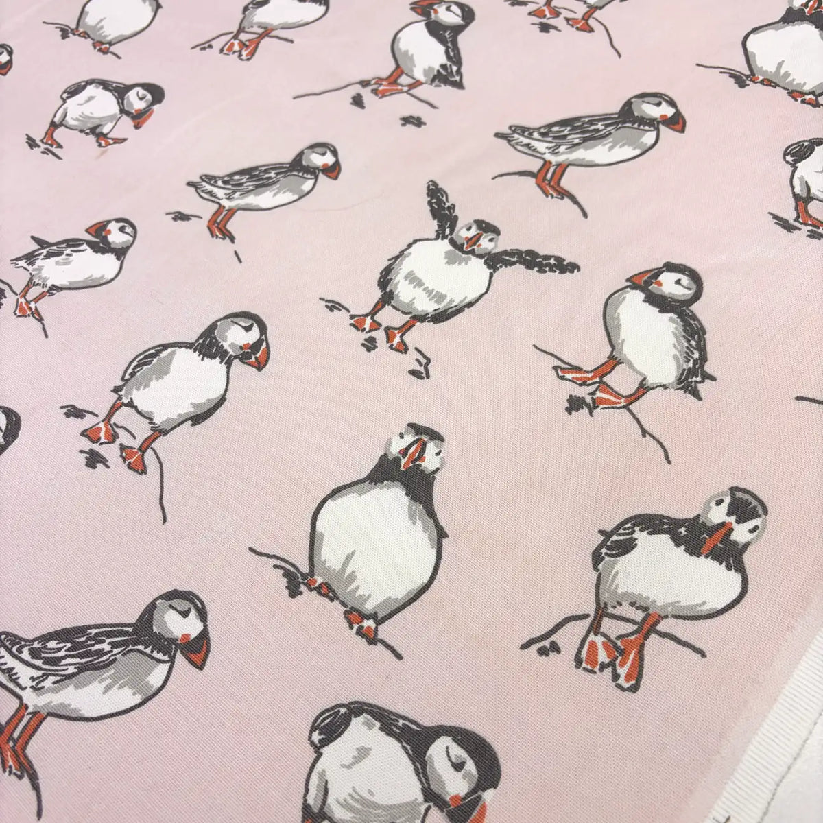 A close-up of a light pink fabric printed with illustrated puffins in black, white, gray, and orange.